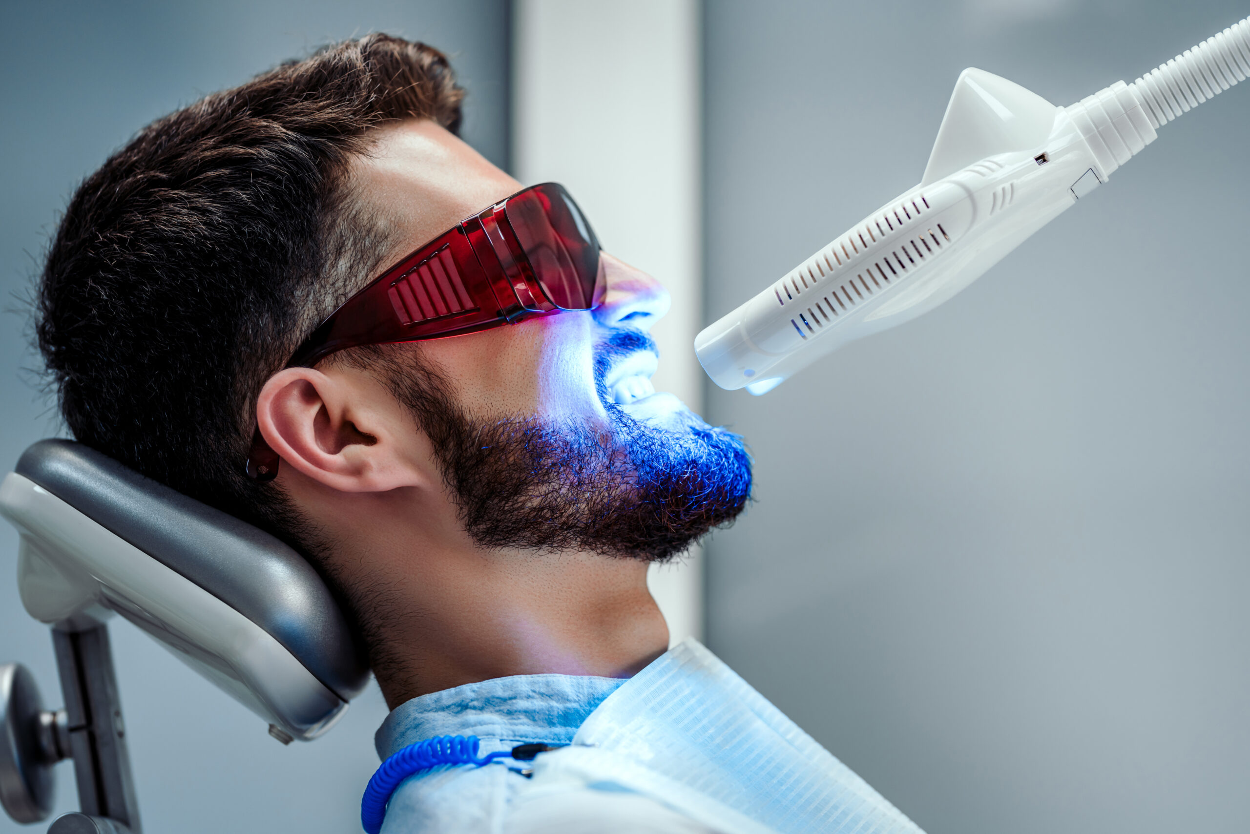 Unrecognizable dentist whitening teeth of young man sitting in dental chair in modern clinic. Side view.