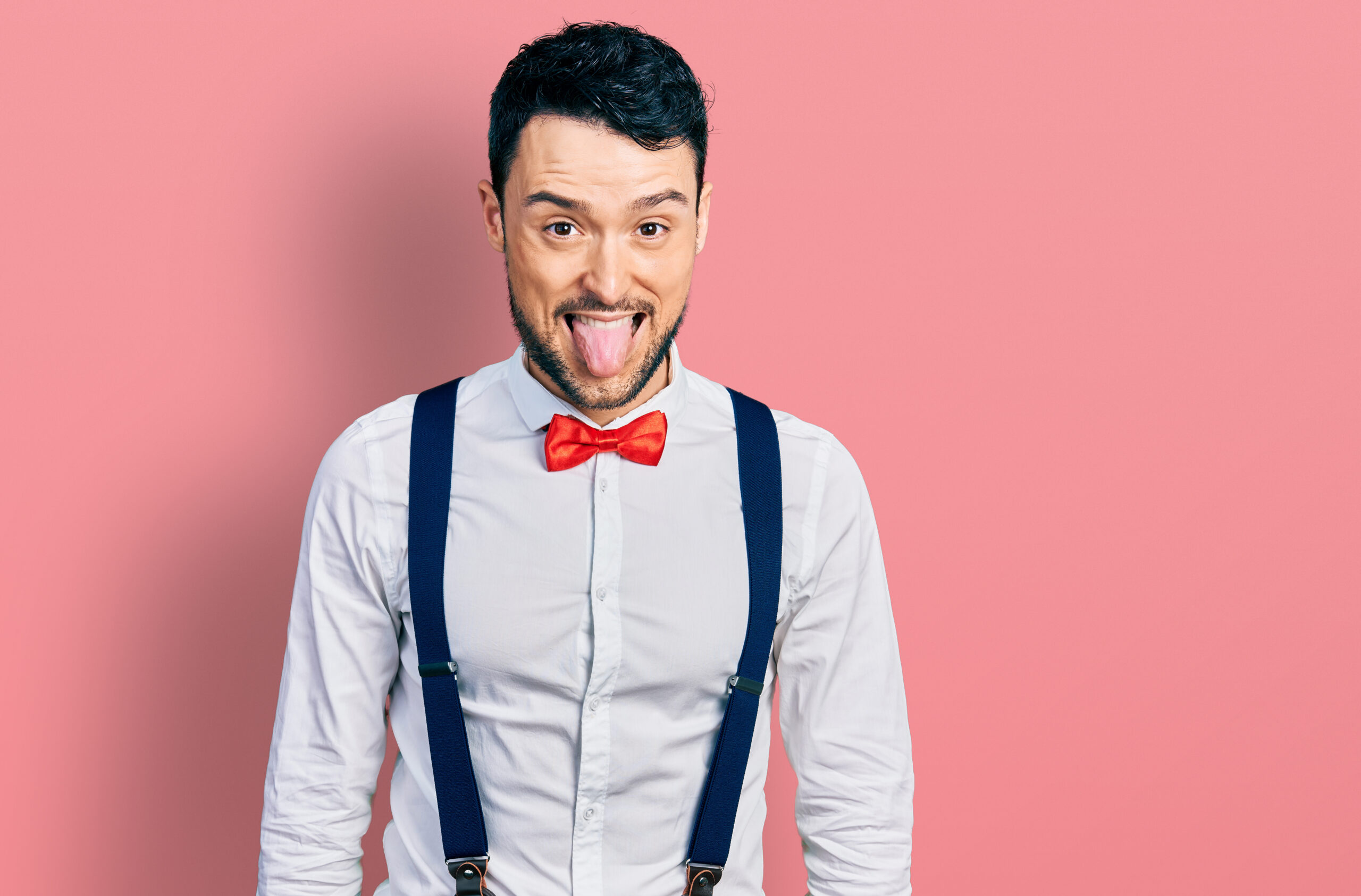 Hispanic man with beard wearing hipster look with bow tie and suspenders sticking tongue out happy with funny expression. emotion concept.